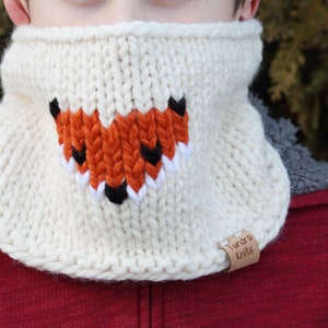 May include: White knitted neck warmer with a fox face design in orange, black, and white. The neck warmer has a brown leather tag that says "Tundra Knits".
