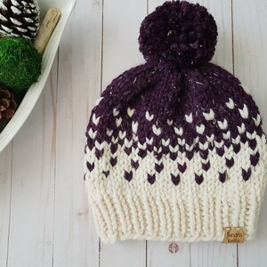 May include: A white and purple knitted beanie with a pom pom on top. The beanie has a white label with the text "Lunda's Knits" on the side.