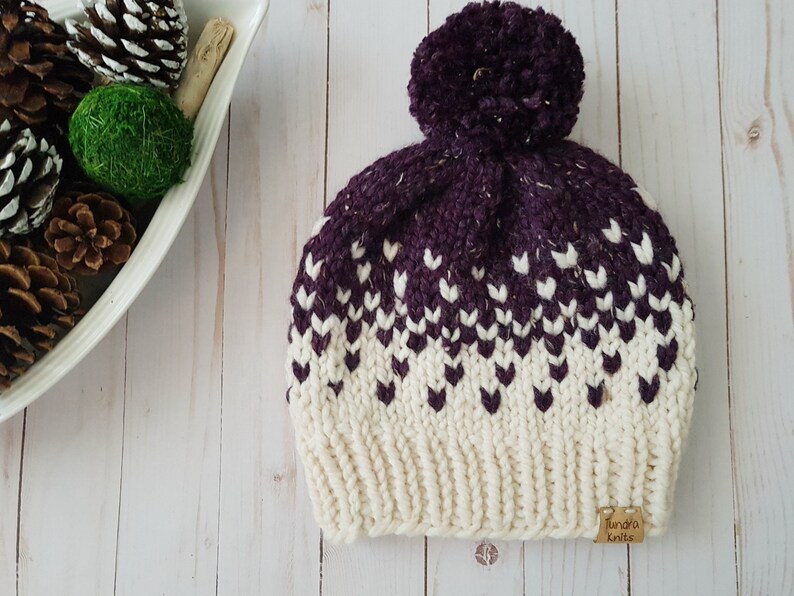 May include: A white and purple knitted beanie with a pom pom on top. The beanie has a white label with the text "Lunda's Knits" on the side.