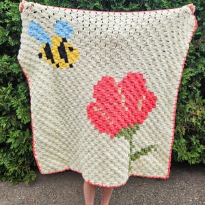 May include: A crocheted baby blanket with a bee and flower design. The blanket is white with a red flower and a yellow bee with blue wings.