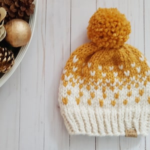 May include: A mustard yellow knit beanie with a white geometric pattern and a matching pom pom. The beanie has a brown leather tag that says "Aurora Knits".