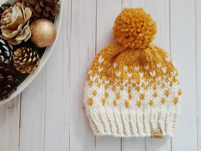 May include: A mustard yellow knit beanie with a white geometric pattern and a matching pom pom. The beanie has a brown leather tag that says "Aurora Knits".