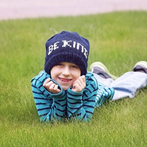 Könnte beinhalten: Ein junges Kind trägt eine blaue Strickmütze mit den Worten "Be Kind" in weißen Buchstaben und liegt auf dem Bauch in einem grasbewachsenen Feld. Es lächelt und hat die Hände unter dem Kinn.