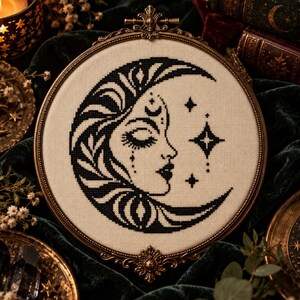May include: A circular embroidery hoop containing a cross-stitch design on off-white fabric. The black thread pattern depicts a crescent moon shaped like a human face in profile, surrounded by small four-pointed stars and decorative accents.