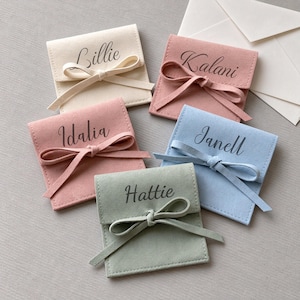 Puede incluir: Cinco bolsitas cuadradas de terciopelo en color crema, rosa viejo, rosa claro, azul claro y verde salvia. Cada bolsita tiene un cierre de lazo a juego y está personalizada con un nombre en letra cursiva oscura: Lillie, Kalani, Idalia, Janell y Hattie.