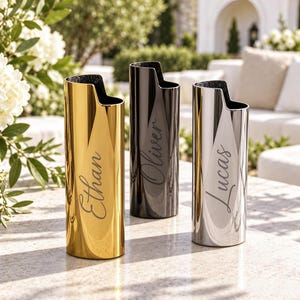 Puede incluir: Tres fundas cilíndricas de metal para encendedores con un borde superior con muescas. La funda izquierda es dorada con el nombre Ethan, la central es negra con Oliver y la derecha es plateada con Lucas, grabadas en letra cursiva.