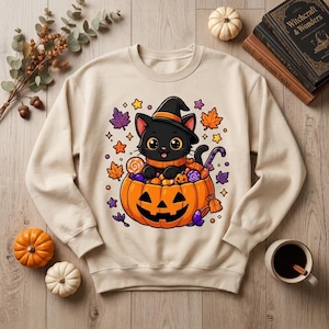 Puede incluir: Una sudadera beige de manga larga con un gráfico de un gato negro con sombrero de bruja, sentado dentro de una calabaza de Halloween tallada llena de dulces, rodeado de hojas de otoño y pequeñas estrellas.
