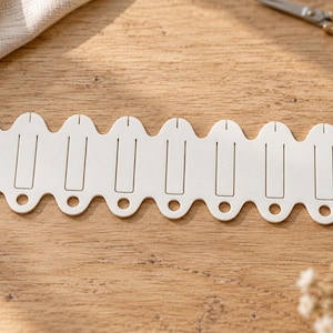 May include: A white, flat, elongated organizer tool featuring a series of seven scalloped sections, each with a vertical rectangular cutout and a circular hole at the base, ending with a single small hole on the right side.