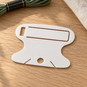 May include: A white, flat, spool-shaped plastic embroidery floss organizer featuring a rectangular cutout at the top, a circular hole at the bottom, and two small angled slits at the base for securing thread ends.