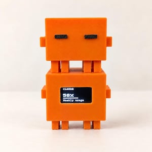 May include: A bright orange, blocky, robot-shaped device with two stacked square sections. The top section features two small black horizontal rectangles for eyes. The bottom section contains a digital screen displaying the text CLAUDE, 58%, and Weekly usage.