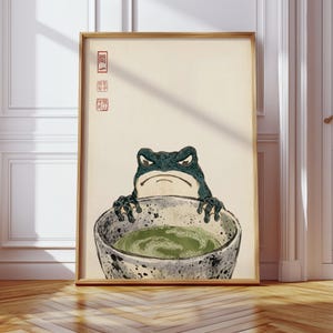 May include: A framed wall art print featuring a dark green frog with a stern expression holding a speckled ceramic bowl filled with green matcha tea. The background is a light cream color with red Japanese characters printed in the upper left corner.