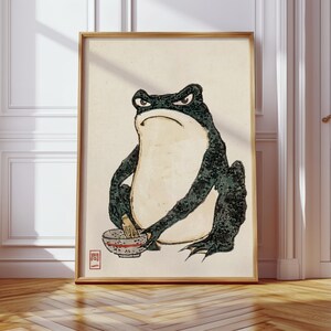 Pode incluir: Uma impressão de arte emoldurada apresentando um sapo verde-escuro com aparência rabugenta e barriga creme. O sapo segura uma pequena tigela com um batedor dentro. No canto inferior esquerdo, há um selo vermelho com o texto japonês 問一.