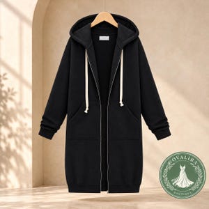 Puede incluir: Una sudadera larga negra con cremallera, capucha con cordones blancos gruesos, mangas largas con puños acanalados y dos bolsillos delanteros. La prenda se muestra en una percha de madera y cuenta con una cremallera frontal completa.