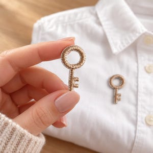 May include: A small, embroidered key-shaped patch featuring metallic gold thread with a white border. The patch has a circular head and a short, notched shaft. A second identical key patch is attached to a white collared shirt.