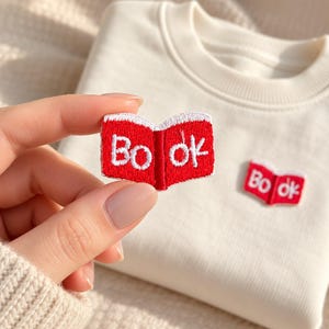 May include: A cream-colored sweatshirt featuring a small, embroidered patch shaped like an open red book with white edges. The word "Book" is stitched in white letters across the two pages of the open book design, which is also visible on the fabric.