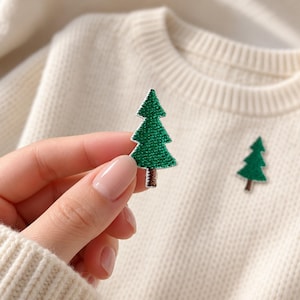 May include: A small, embroidered patch shaped like a green pine tree with a brown trunk. The patch features a textured, stitched surface and is shown against a cream-colored, ribbed knit sweater that has an identical tree patch attached.