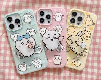 May include: Three smartphone cases in light blue, pink, and yellow, each featuring a matching character phone grip and printed cartoon illustrations of small white creatures. The cases have camera cutouts and the text Chiikawa printed at the bottom.