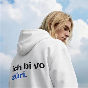 Puede incluir: Una sudadera con capucha blanca que presenta el texto negro 'ich bi vo' y el texto azul 'züri.' impresos en la parte posterior. La prenda tiene capucha y mangas largas, vista desde un ángulo trasero con un cielo nublado.