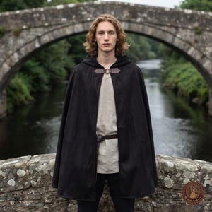 May include: A black hooded cloak featuring a brown leather neck closure with a metal ring. The cloak is worn over a light beige tunic and a dark brown leather belt. The garment is solid black with a smooth texture and a hood.