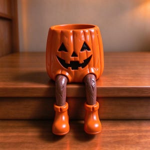 Puede incluir: Una maceta de cerámica naranja brillante con forma de calabaza y un rostro tallado de jack-o'-lantern con ojos triangulares y una amplia sonrisa. Tiene dos piernas marrones texturizadas que terminan en botas naranjas lisas.