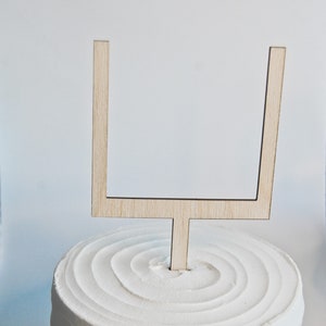 May include: A white cake with a wooden football goalpost topper. The cake is decorated with a swirl pattern and the words "Kendol Made" are written on the bottom.