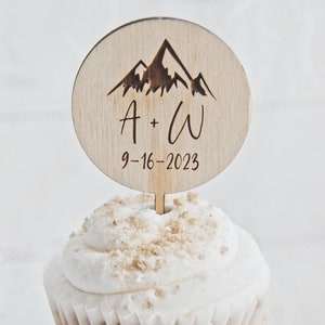 May include: A wooden cupcake topper with a mountain range design and the initials "A + W" and the date "9-16-2023".