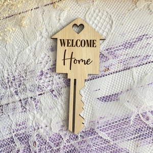 May include: Wooden key-shaped home decor with a heart cutout and the words "WELCOME Home" engraved on it. The key is on a white lace background.