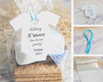 May include: A clear plastic bag filled with graham crackers, marshmallows, and a white chocolate square. A white tag with a baby onesie shape is attached to the bag with blue and white twine. The tag reads "Adding S'more love to our family! Nolan 2023".