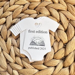 May include: A white baby onesie with a black and white illustration of a book with a sunburst above it. The text "first edition" and "published 2025" is printed on the onesie.