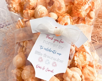 Puede incluir: Una bolsa de plástico transparente llena de palomitas de maíz con caramelo con una etiqueta blanca adjunta. La etiqueta tiene forma de mono y tiene el texto "Thank you for POPPIN' BY to celebrate our little firecracker Dan & Sarah" con ilustraciones de fuegos artificiales rojos, blancos y azules.