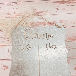 May include: A galvanized metal sign with a decorative edge and a jute rope hanger. The sign features the word "Oihana" in wooden script, along with "To Do" and "Done". A vertical wooden strip divides the sign.