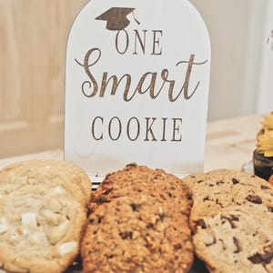 May include: A graduation-themed display featuring three large cookies on a white cake stand. A wooden sign with a graduation cap graphic reads "ONE Smart COOKIE". The cookies are various shades of brown.