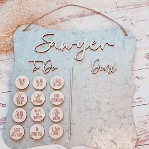 May include: A galvanized metal chore chart with the name "Sawyer" in wooden script. The chart has a "To Do" and "Done" section, with wooden circles listing chores like "Make Bed" and "Clean Closet". A jute rope is attached for hanging.