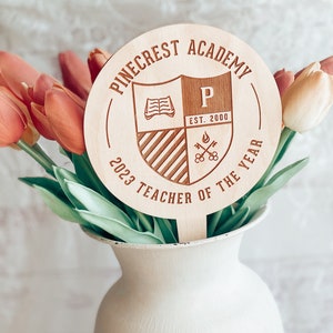 May include: A wooden plaque with the text "Pinecrest Academy" and "2023 Teacher of the Year" around a shield with a book, the letter "P", and the text "Est. 2000".