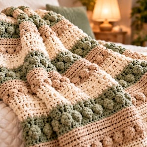 May include: A crocheted blanket featuring alternating horizontal stripes in sage green, cream, and beige. The design incorporates textured bobble stitch rows interspersed with sections of open, airy crochet work, creating a layered appearance.