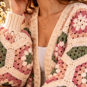 May include: A crochet cardigan featuring a patchwork of hexagonal motifs in cream, sage green, and dusty rose yarn. The garment has a ribbed cream collar and cuffs, a V-neckline, and is worn over a plain white top with a gold pendant necklace.