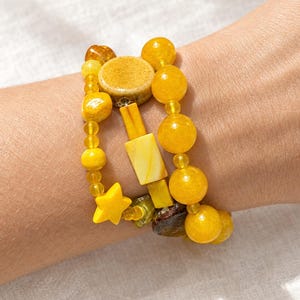 May include: A multi-strand bracelet featuring a variety of yellow beads in different shapes and sizes, including round spheres, rectangular blocks, a flat circular disc, and a five-pointed star charm, all strung together on a flexible cord.