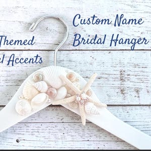 May include: A white wooden clothes hanger featuring a silver metal hook and a collection of various seashells, a starfish, and small clear jewel accents attached to the top. The text Custom Name Bridal Hanger and Beach Themed w/Jewel Accents is visible.