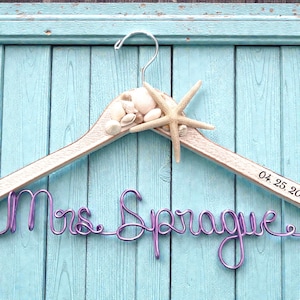 May include: A wooden hanger decorated with seashells and a starfish. The hanger features the name "Mrs. Sprague" in purple wire and the date "04. 25. 20" printed on the wood. The background is a light blue wooden surface.