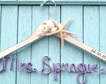 May include: A wooden hanger decorated with seashells and a starfish. The hanger features the name "Mrs. Sprague" in purple wire and the date "04. 25. 20" printed on the wood. The background is a light blue wooden surface.