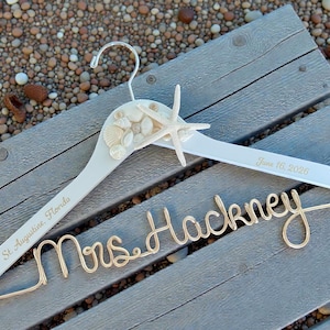 May include: A white wooden wedding dress hanger decorated with seashells and a starfish. The hanger is personalized with the text "St. Augustine, Florida" and "June 16, 2026". A gold wire name "Mrs. Hackney" is attached.