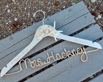 May include: A white wooden wedding dress hanger decorated with seashells and a starfish. The hanger is personalized with the text "St. Augustine, Florida" and "June 16, 2026". A gold wire name "Mrs. Hackney" is attached.