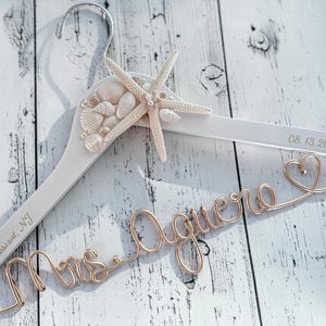 May include: A white wooden clothes hanger featuring a rose gold wire script reading Mrs. Aguero with a heart shape. The hanger is decorated with a cluster of white seashells and a starfish. Engraved text reads Point Pleasant, NJ and 08.13.26.