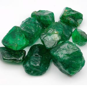 May include: A collection of raw, unpolished emerald gemstones with a deep green color and irregular, jagged shapes. The stones feature a translucent to opaque appearance with natural surface textures and varying sizes clustered together.
