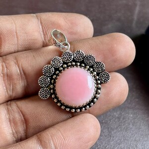 Könnte beinhalten: Ein runder Anhänger mit einem glatten, hellrosa Cabochon-Stein, der in einen dekorativen silberfarbenen Metallrahmen gefasst ist. Der Rahmen hat einen gewellten Rand mit kleinen, erhabenen kreisförmigen Texturen und eine einfache Öse.