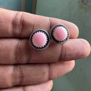 Könnte beinhalten: Ein Paar runde Ohrstecker mit glatten, hellrosa Cabochon-Steinen, die in texturierte, oxidierte Sterlingsilberfassungen mit einem dekorativen seilartigen Randdetail um die kreisförmige Fassung herum eingefasst sind.