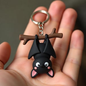 May include: A black bat keychain featuring a matte finish, hanging upside down from a brown branch. The bat has large white eyes with black pupils, small pink inner ears, and pink circular cheek accents, attached to a silver metal split ring.