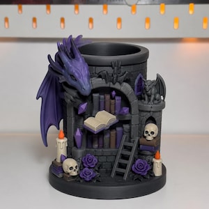 May include: A cylindrical dark gray desk organizer featuring a purple dragon, a stone tower facade with shelves of books, an open book, a ladder, two skulls, two candles with orange flames, purple roses, and a small gargoyle figure.