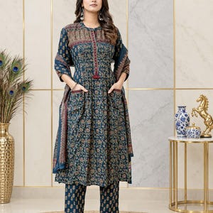 May include: A dark teal long-sleeved kurta dress featuring a repeating floral pattern in gold and red, a buttoned placket with a small tassel, two front pockets, and matching straight-leg trousers, accompanied by a coordinating printed shawl.