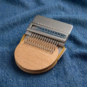 May include: A darning loom tool featuring a circular light-colored wooden base, a rectangular metal plate with a series of vertical hooks, and a set of blue metal clips secured across the top section of the device for textile repair.